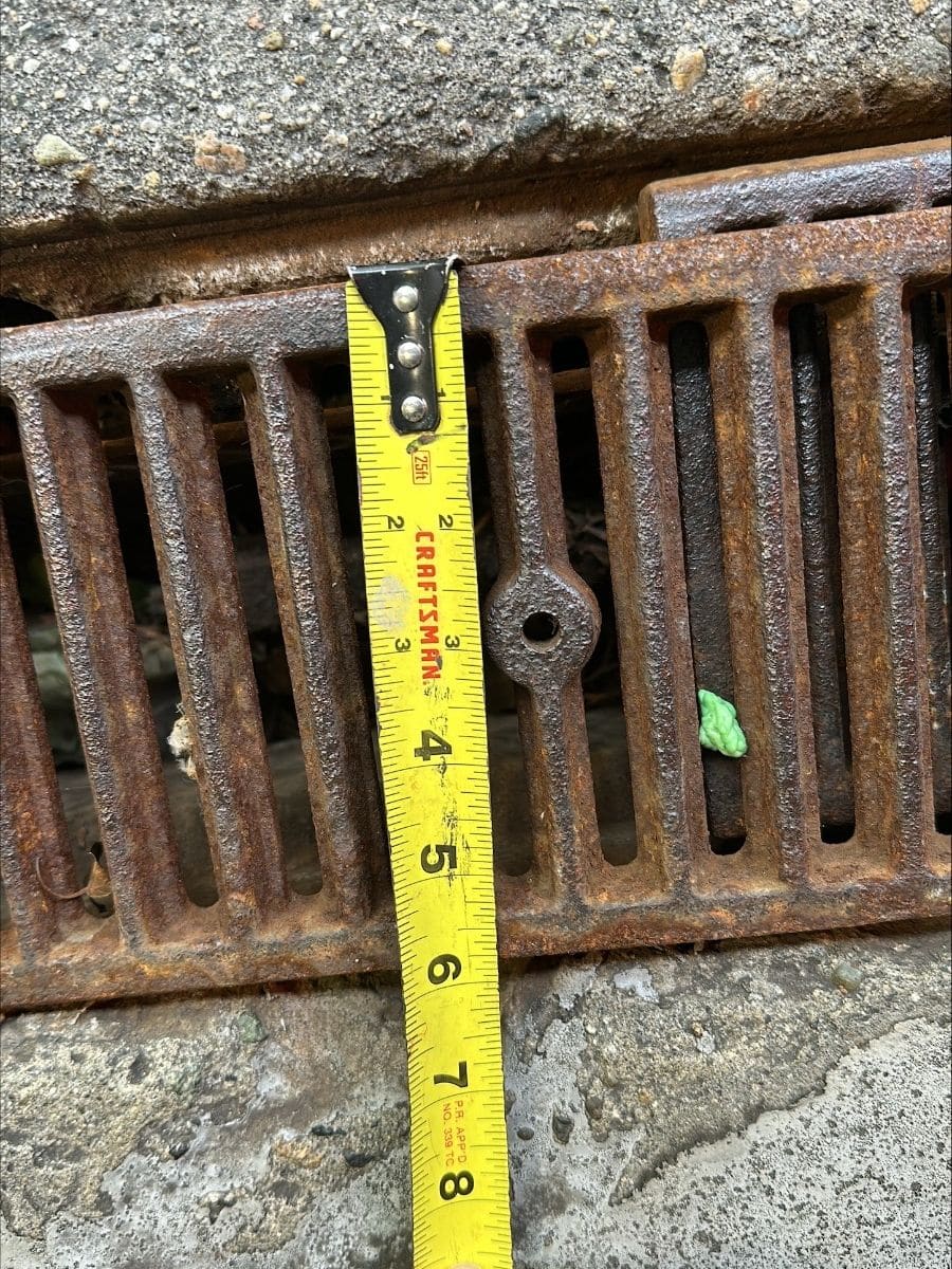 A tape measure held across a rusted trench drain grate set in concrete